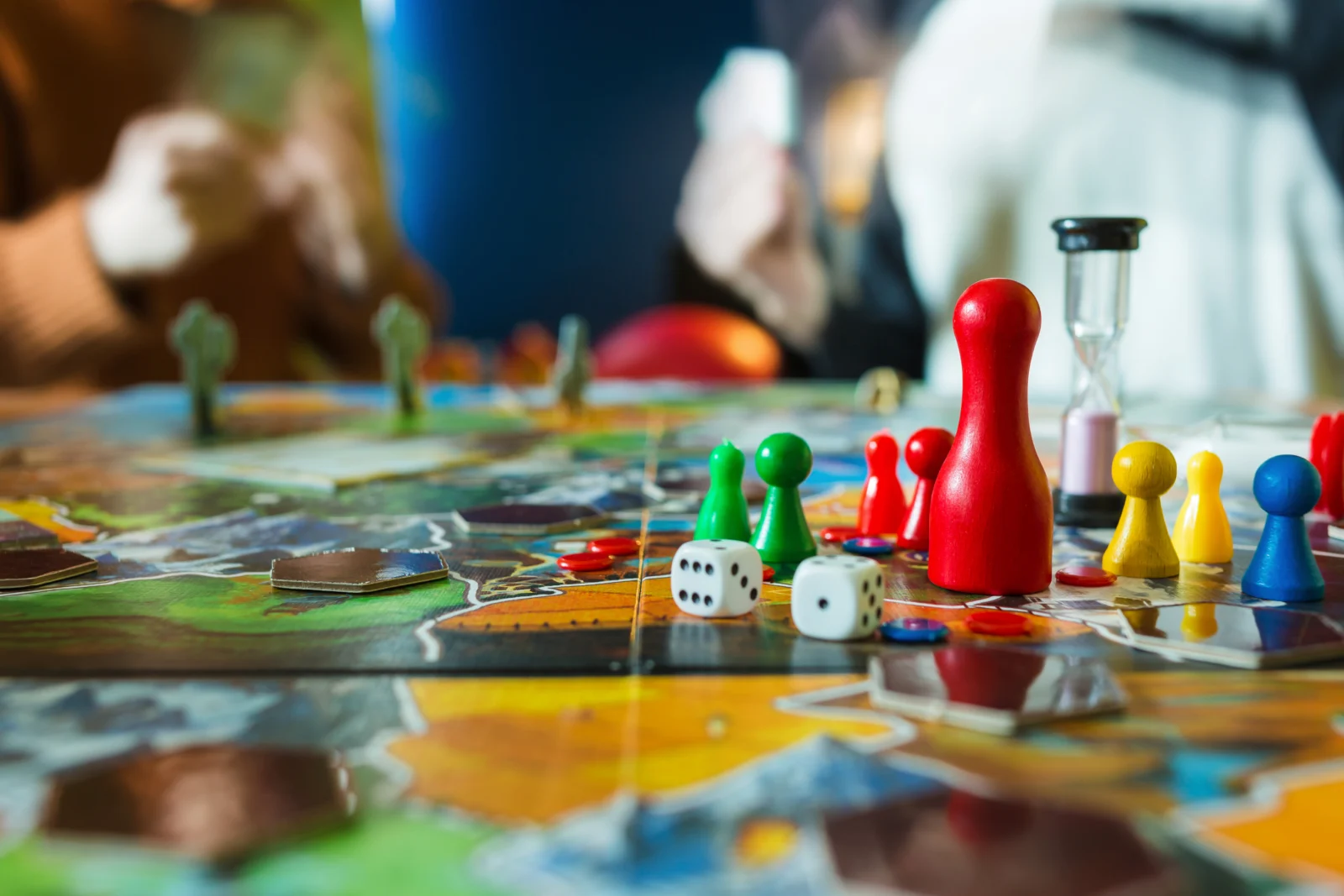 Spielbrett mit bunten Spielfiguren, Würfeln und einer Sanduhr, im Hintergrund zwei Personen mit Spielkarten.