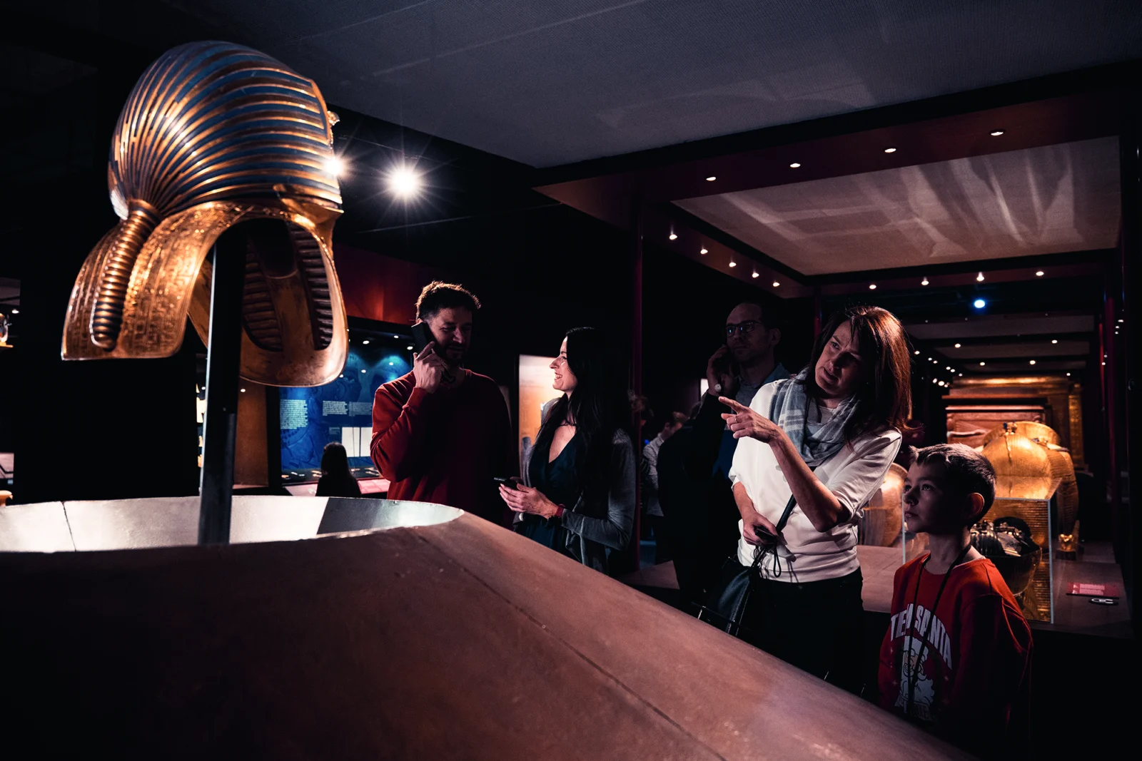 Ausstellungsraum mit einer goldenen Pharaonenmaske im Vordergrund, dahinter Besucherinnen und Besucher mit einem Kind.